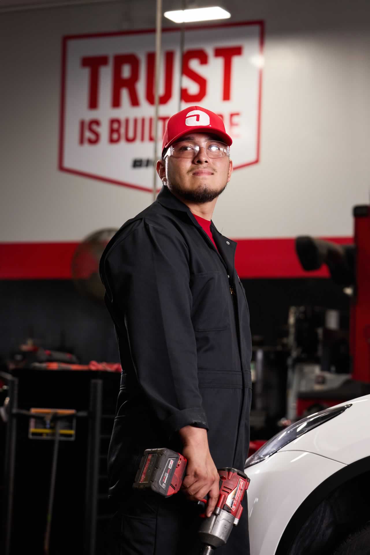 Mechanic in a workshop stands near a vehicle holding a power tool, wearing a red cap and black coveralls. A sign in the background reads "TRUST IS BUILT.