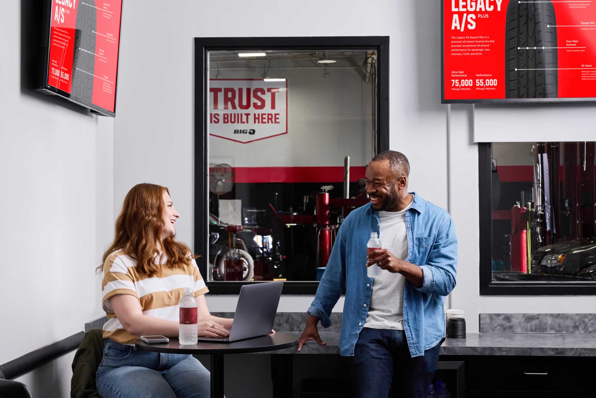 Two people converse in an auto shop waiting area. One sits with a laptop, and the other stands holding a drink. Wall signs display tire information.