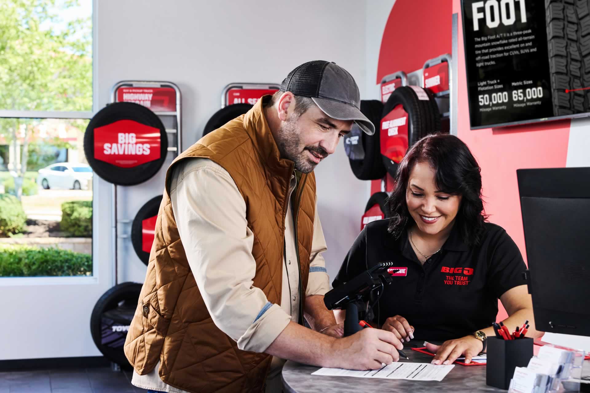 A man signs a document at a store counter while a woman in a uniform smiles beside him. Tire displays and a sign with promotions are visible in the background.