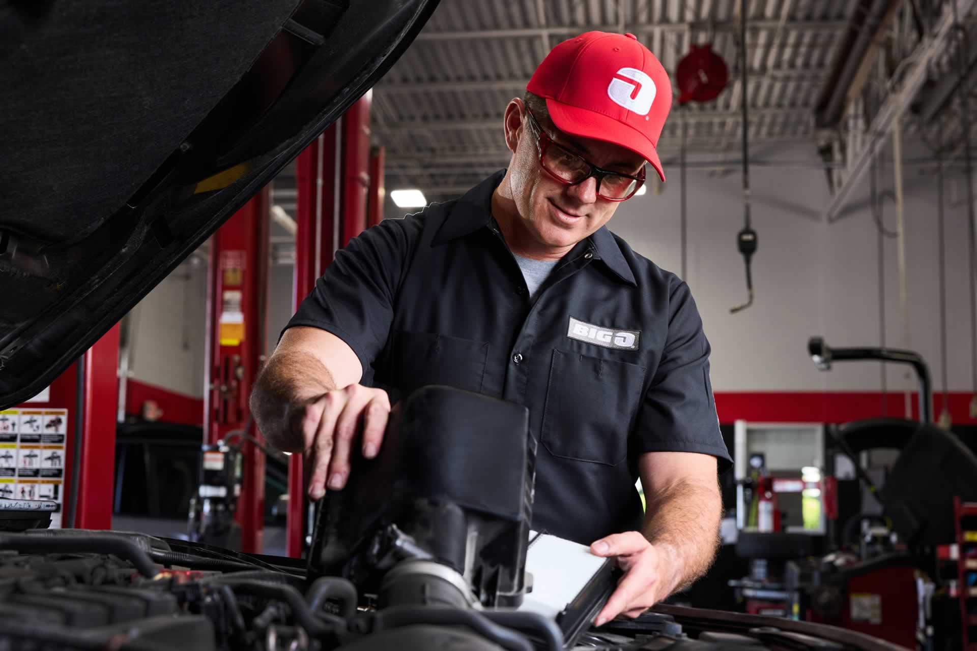 Mechanic in a red hat and black uniform inspects an engine in an auto repair shop.