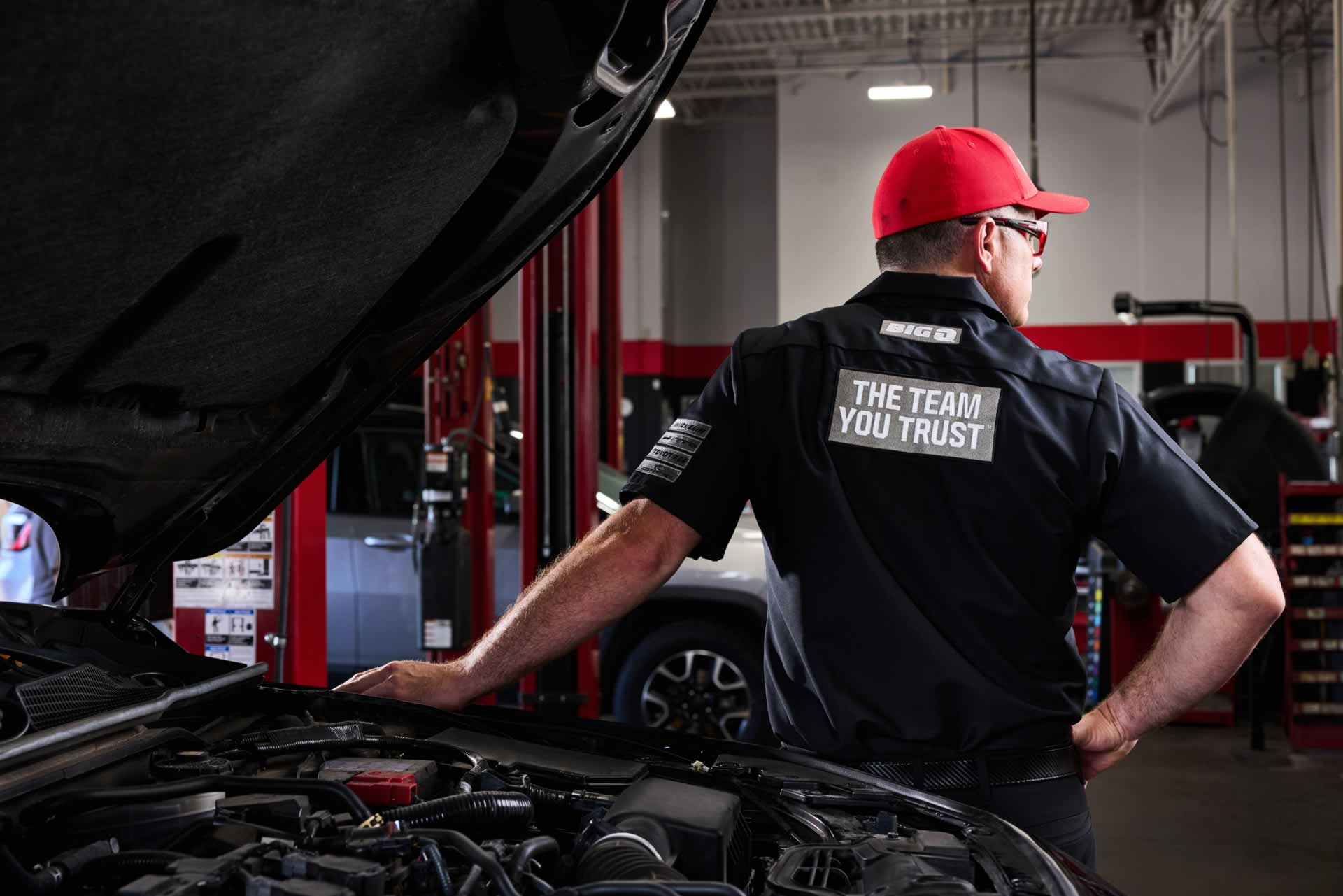 Mechanic in a red cap and black uniform with "The Team You Trust" on the back, stands by a car with the hood open in an auto repair shop.