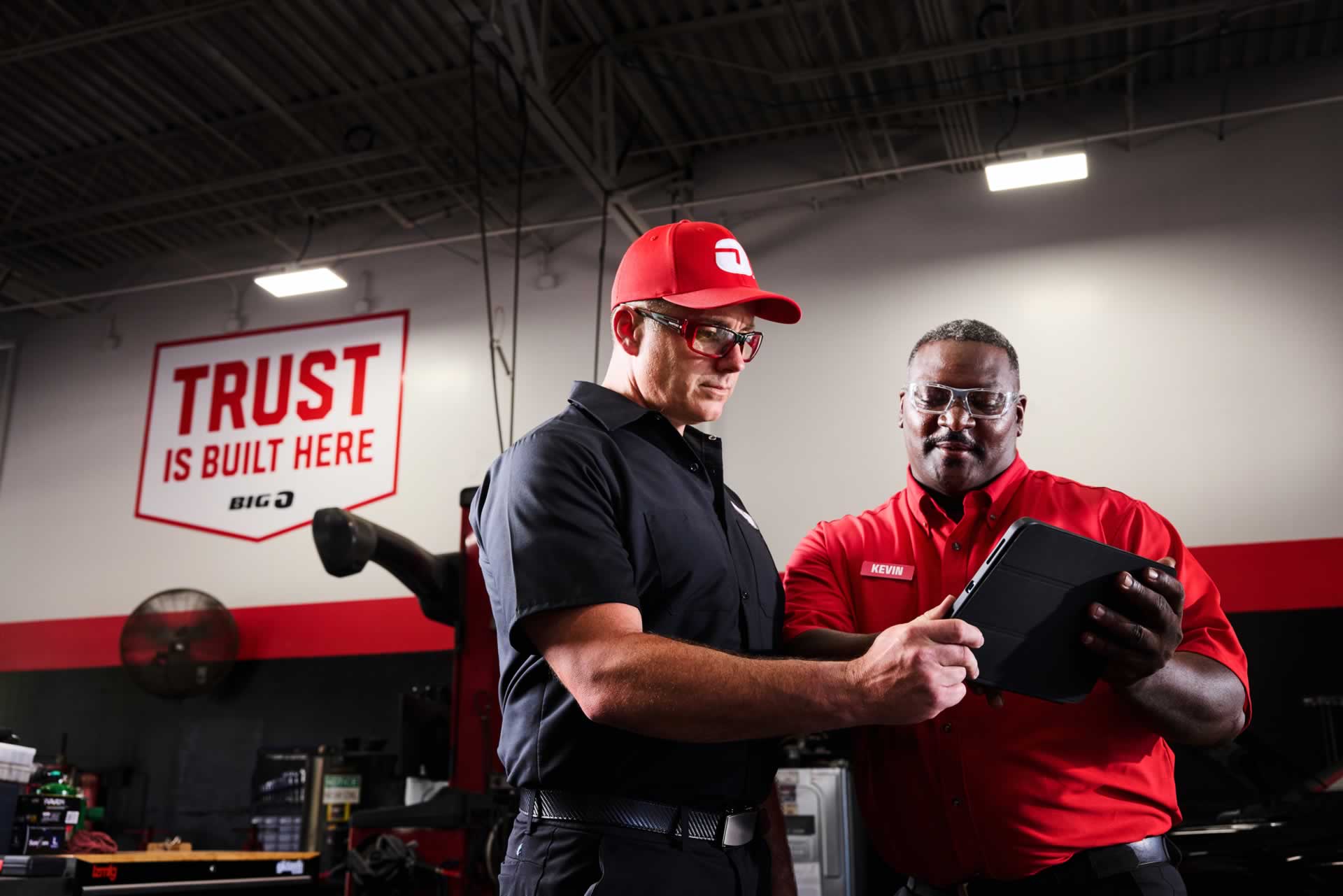 Two mechanics in a garage review information on a tablet. A sign on the wall reads "TRUST IS BUILT HERE.