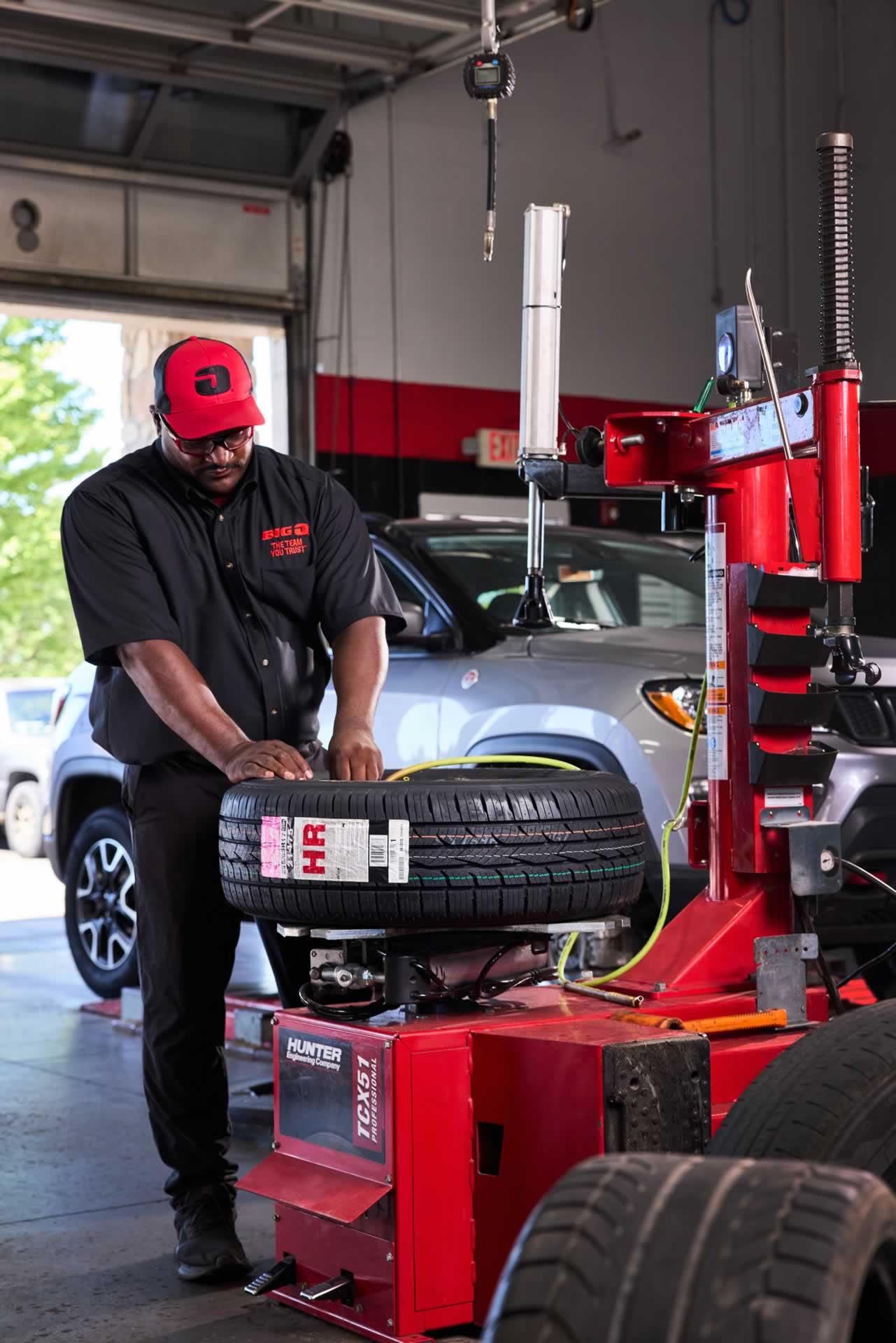 A mechanic in a black uniform operates a tire mounting machine in an auto shop, with a car visible in the background.