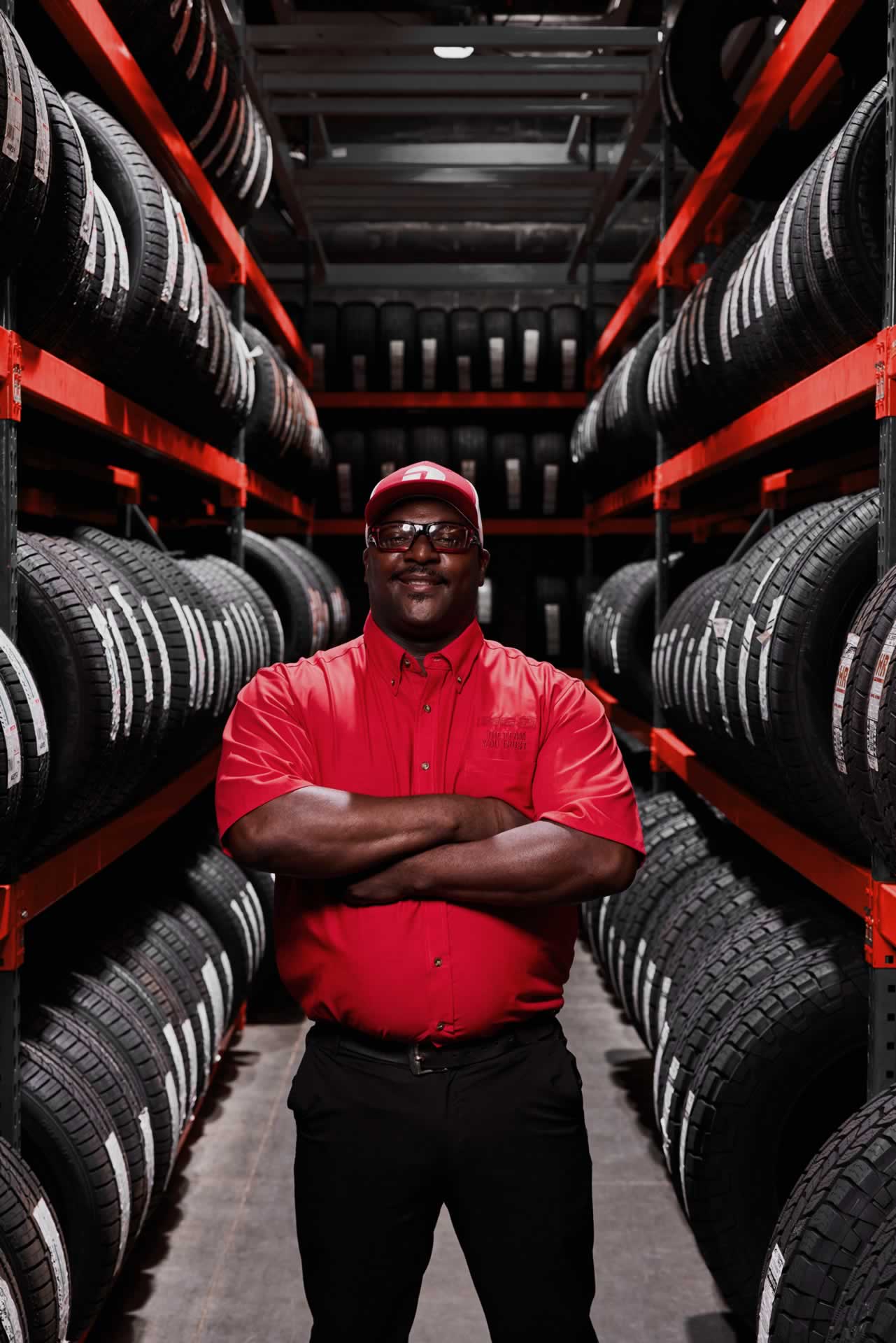 A person in a red uniform and cap stands with arms crossed in a tire warehouse, surrounded by rows of stacked tires.