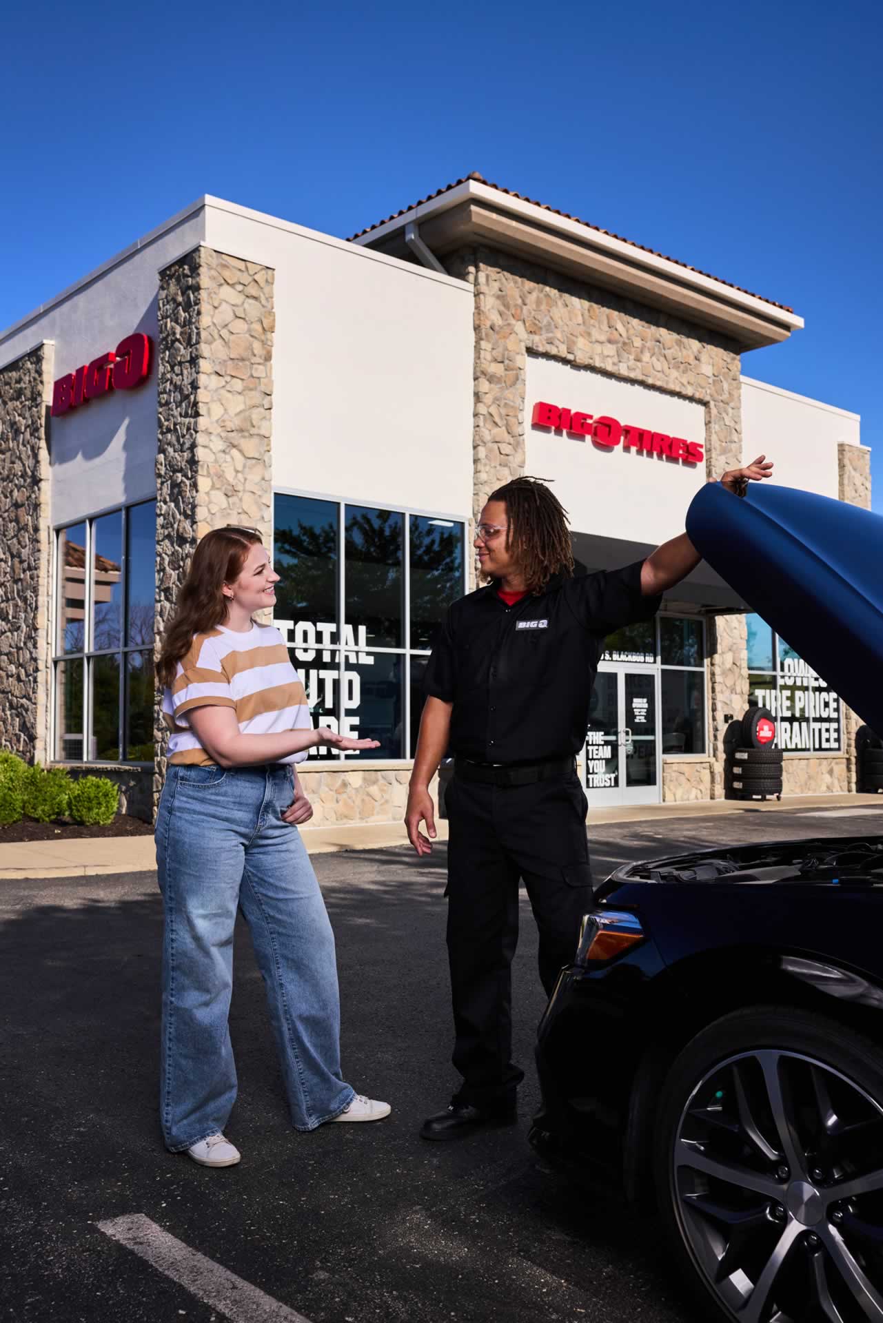 A mechanic gestures while speaking to a woman next to a car with an open hood in front of a tire service store.