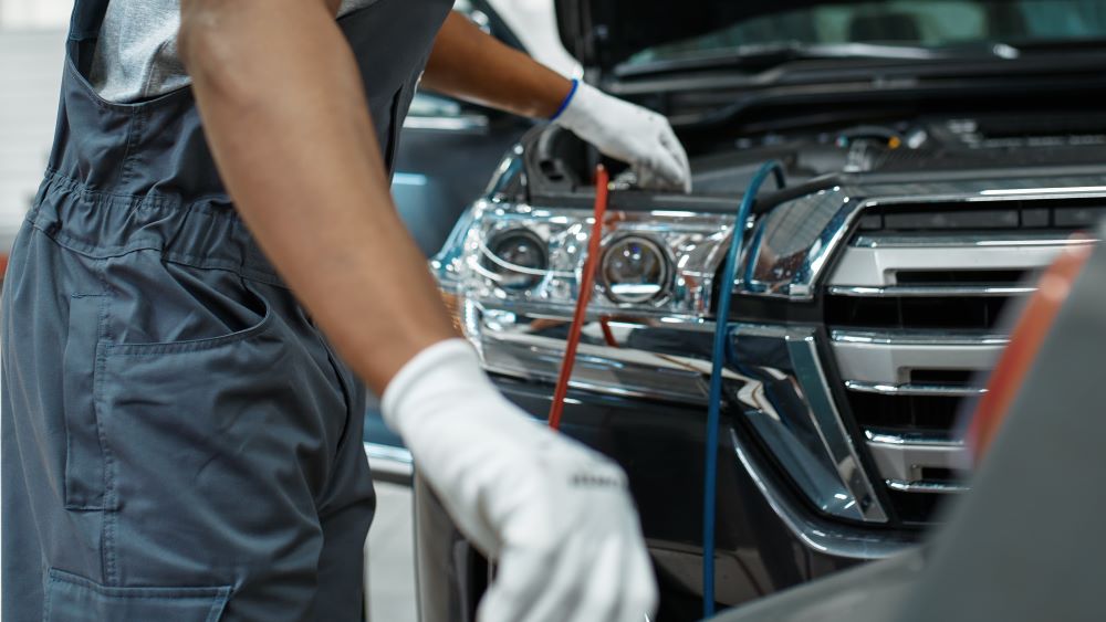 A person in a mechanic's uniform and gloves is working on a car with the hood open at a wheel and tire franchise.